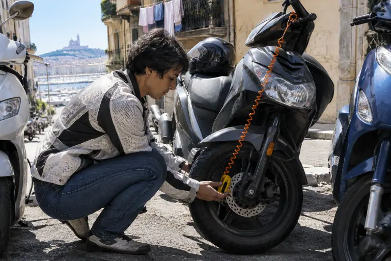Une motarde pose un bloque-disque jaune sur le disque avant de son scooter 125, le câble de rappel orange relié au levier de frein, à Marseille