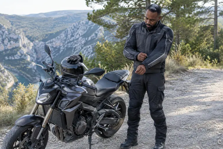 Un motard serre la patte de réglage de l’avant-bras de son blouson textile gris, à côté de son roadster garé au bord d’une route de gorges en Provence