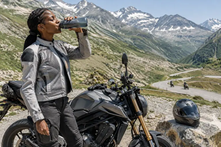 Une motarde en blouson mesh gris clair boit une gorgée d’eau à côté de son roadster, arrêtée au sommet d’un col alpin en plein mois de juillet