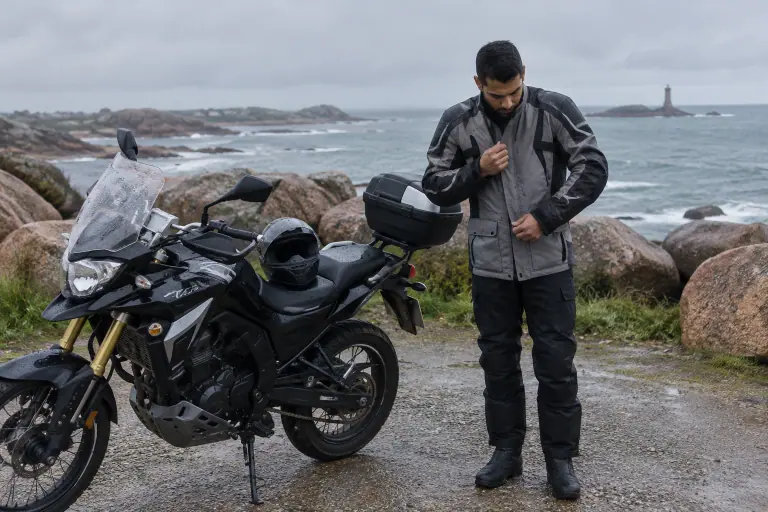 Un jeune motard ferme le rabat de son blouson textile trois quarts perlé de pluie, à côté de son trail garé face aux rochers de la côte bretonne