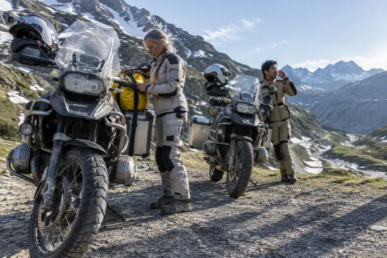 Au sommet d’un col savoyard au petit matin, une motarde sangle son sac étanche sur un gros trail à moteur boxer pendant que son compagnon boit un café