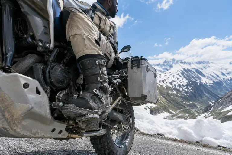 Au sommet d’un col alpin, un motard assis sur un gros trail glisse la pointe de sa botte adventure à boucles sous le sélecteur de vitesses