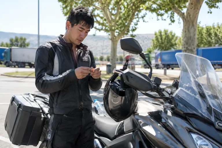Sur une aire d’autoroute en été, un motard roule un bouchon d’oreille en mousse entre ses doigts à côté de sa routière, casque intégral accroché au rétroviseur
