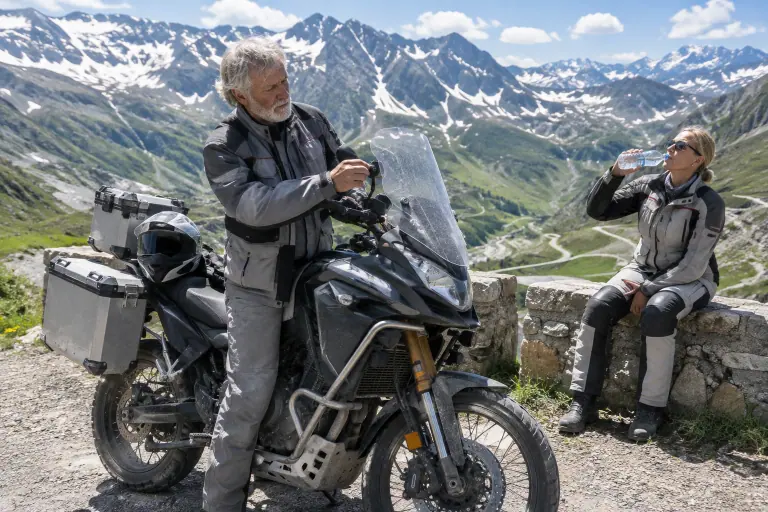 Sur un col alpin en été, un motard à barbe blanche règle la hauteur de la bulle de son gros trail chargé de valises, sa compagne assise à côté