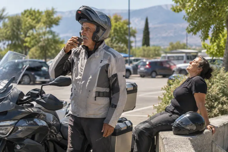 Sur une aire d’autoroute en été, un motard boit un café, la mentonnière de son casque modulable relevée, à côté de sa routière et de sa passagère