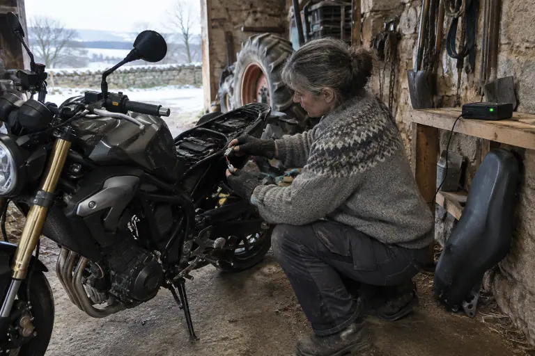 Dans une grange d’Auvergne en hiver, une motarde branche un petit chargeur mainteneur sur le câble à œillets de la batterie de son roadster