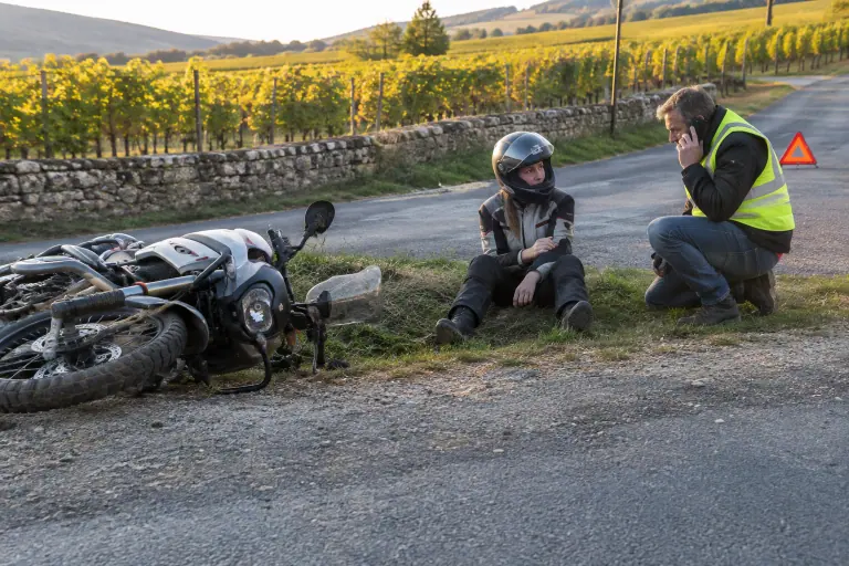 Après une chute sans gravité près des vignes de Bourgogne, une motarde assise sur le bas-côté garde son casque pendant que son ami en gilet jaune appelle