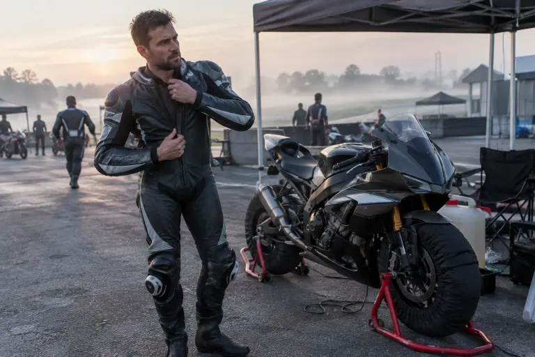 Dans un paddock au petit matin, un motard ferme sa combinaison cuir une pièce près de sa sportive posée sur béquilles d’atelier