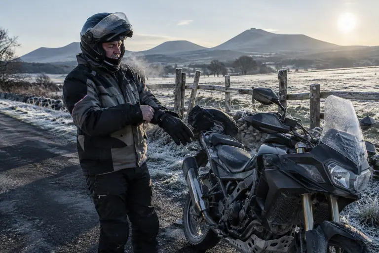 Un motard d’une soixantaine d’années enfile ses gants d’hiver près de son trail équipé de protège-mains, sur une route givrée du plateau auvergnat