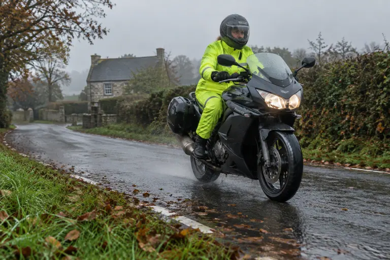 Une motarde en combinaison de pluie jaune fluo roule sur une petite route bretonne détrempée, entre les haies, feuilles mortes et marquages blancs luisants