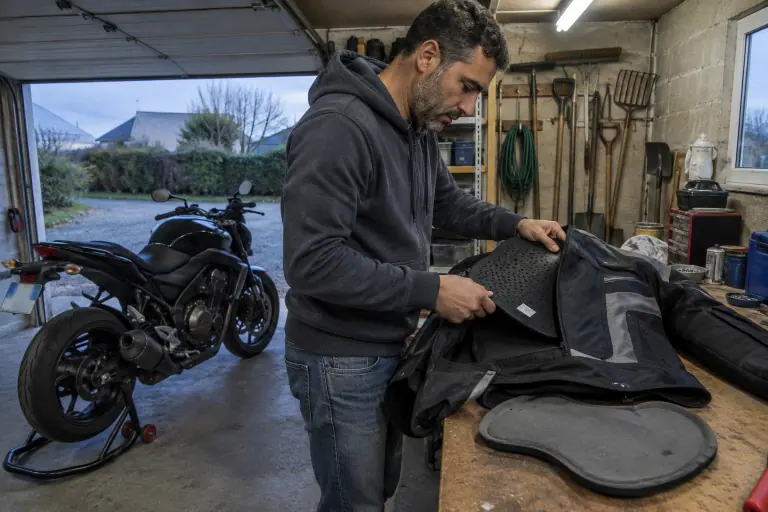 Dans un garage de pavillon, un motard glisse une dorsale perforée dans la poche du dos de son blouson textile, à côté de l’ancienne mousse retirée