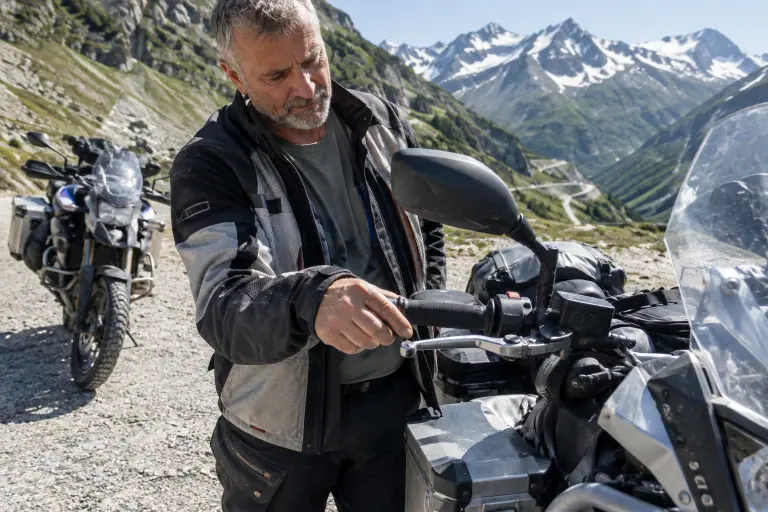 Un motard d’une cinquantaine d’années règle la molette du levier d’embrayage de son trail chargé, sur un parking de gravier près d’un col alpin
