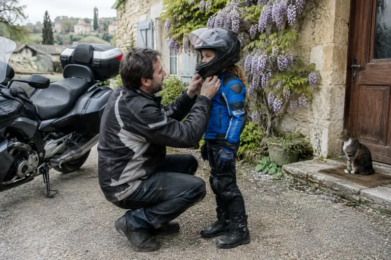 Devant une maison de pierre en Dordogne, un père ferme la jugulaire du casque intégral de sa fille de dix ans, équipée d’un blouson, de gants et de bottes moto