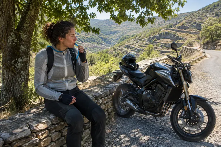 Sur une petite route des Cévennes en juillet, une motarde en blouson mesh boit au tuyau de sa poche à eau, assise à l’ombre d’un châtaignier près de sa moto