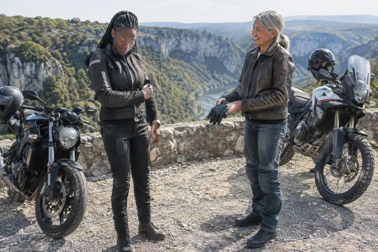 Au belvédère des gorges de l’Ardèche, deux motardes d’âges différents ajustent leur équipement près d’un roadster et d’un trail