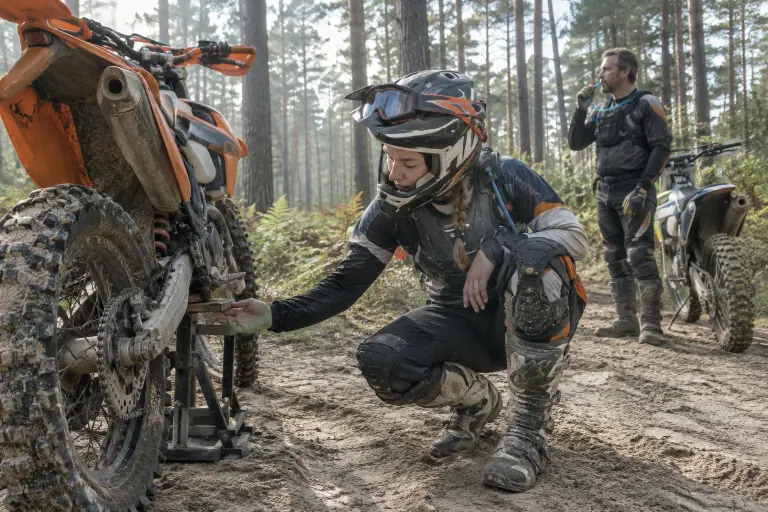 Dans une forêt de pins des Landes, une enduriste vérifie la tension de chaîne de sa moto orange posée sur un lève-moto, au bord d’une piste sableuse