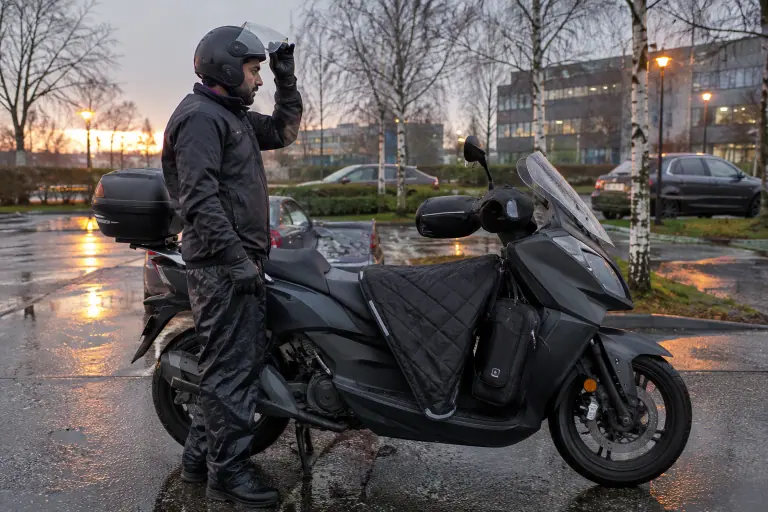 À l’aube de novembre près de Lille, un homme relève la visière de son casque jet à côté d’un scooter 125 équipé d’un tablier, de manchons et d’un top case