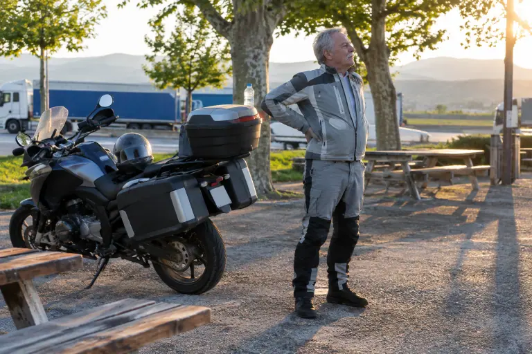 Sur une aire d’autoroute au petit matin, un motard d’une soixantaine d’années s’étire le dos à côté de sa routière chargée de valises et d’un top case
