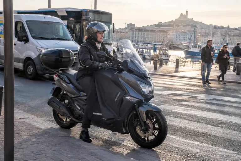 Une femme en blouson textile et casque modulable attend au passage piéton sur un maxi-scooter avec tablier et top case, près du Vieux-Port de Marseille