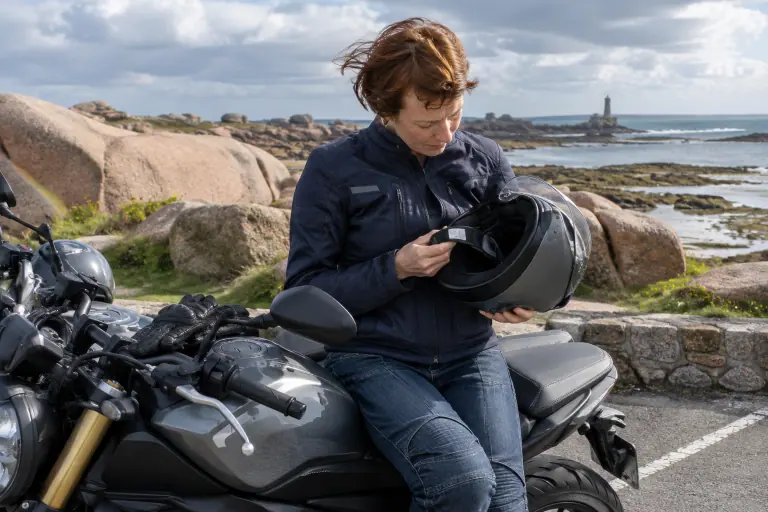 Sur un parking du bord de mer breton, une motarde assise sur son roadster retourne son casque intégral pour lire l’étiquette d’homologation de la jugulaire