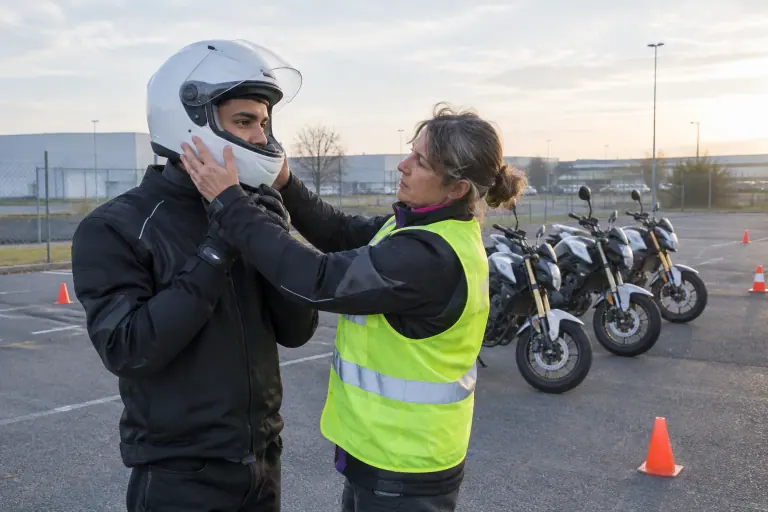 Sur le parking d’une moto-école, une monitrice vérifie le maintien du casque intégral blanc d’entrée de gamme d’un jeune élève avant la leçon