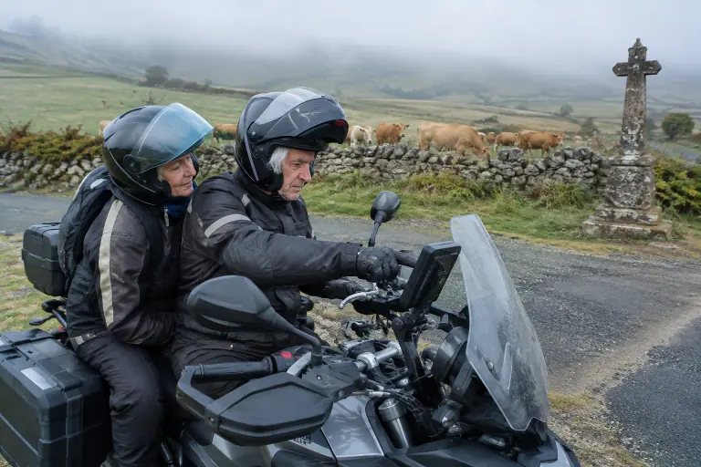 Sur le plateau de l’Aubrac dans la brume, un motard retraité montre à sa passagère l’écran du GPS fixé au guidon de leur routière, près d’une croix de granit