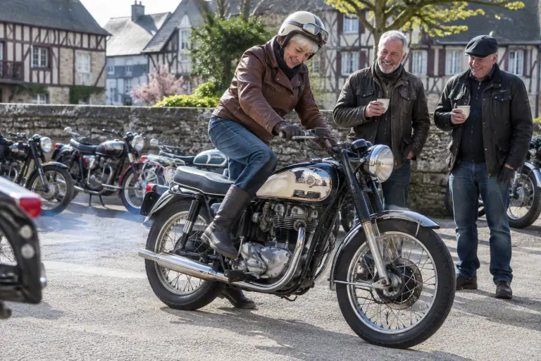 Une motarde aux cheveux blancs démarre au kick une moto ancienne bicylindre lors d’un rassemblement de club sur la place d’un village normand