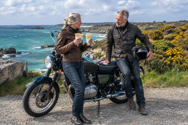 Un couple de motards d’une cinquantaine d’années fait une pause crêpes près d’une moto néo-rétro à phare rond et jantes à rayons, au bord de la mer en Bretagne