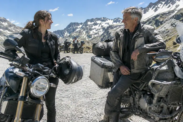Au sommet d’un col alpin, une jeune motarde et un motard plus âgé discutent devant un roadster récent et un vieux trail chargé de valises en aluminium