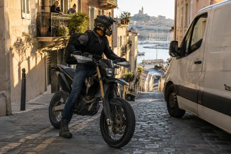 Dans une rue pentue de Marseille, un jeune motard en casque crossover attend sur son supermotard à protège-mains, un pied posé sur les pavés