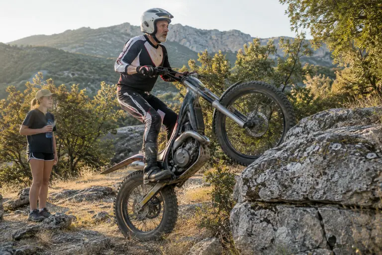Un trialiste d’une soixantaine d’années, debout sur les repose-pieds, hisse sa moto de trial sans selle sur une marche rocheuse, observé par sa petite-fille