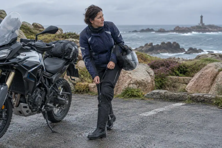 Sur un parking face à la côte de granit bretonne, une motarde remonte la longue fermeture latérale de son surpantalon moto au-dessus de ses bottes
