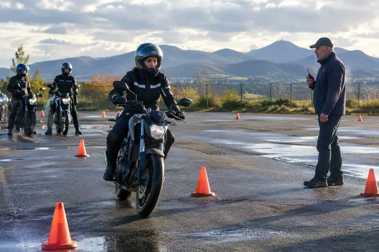Sur la piste d’une moto-école, une élève roule au pas entre des plots orange sous le regard de son moniteur, radio en main, un matin d’automne