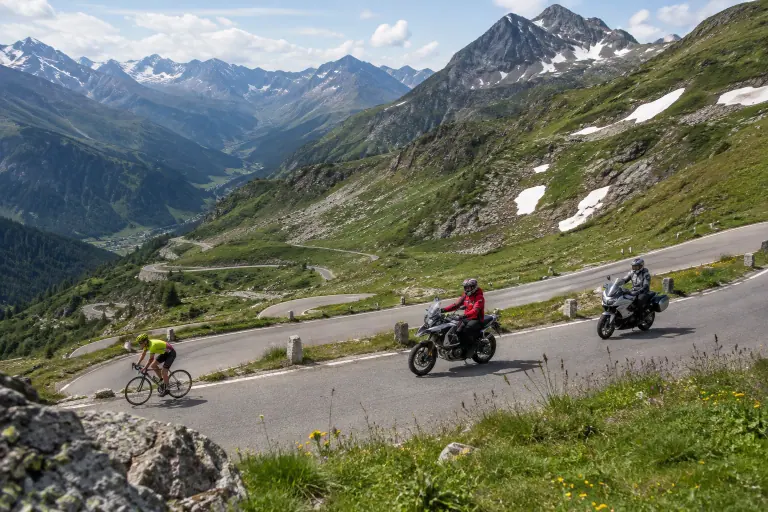 Deux motos montent tranquillement les lacets d’un col alpin en juillet, à côté d’un cycliste, entre alpages fleuris et névés