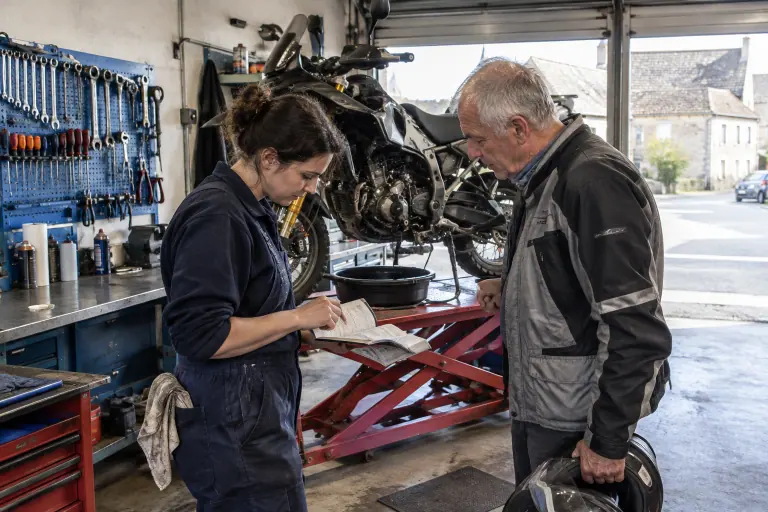 Dans un petit atelier de Bourgogne, une mécanicienne parcourt le carnet d’entretien avec un motard, devant un gros trail levé sur un pont