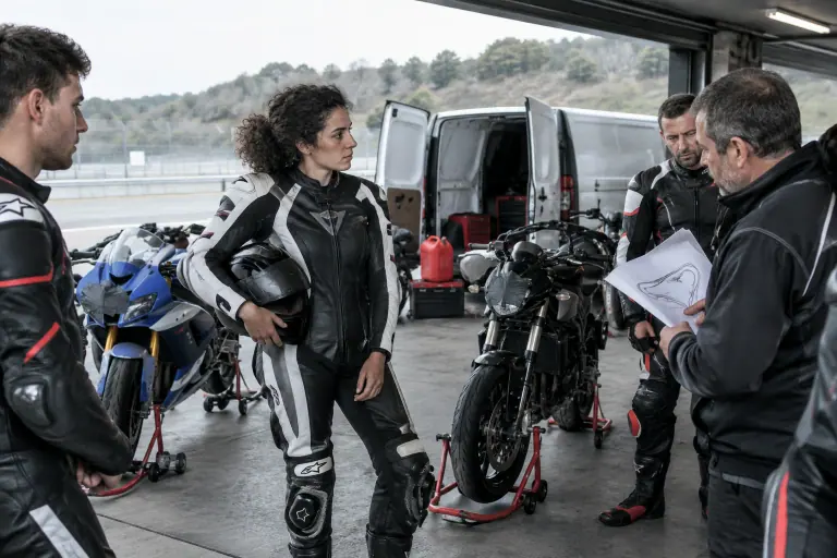 Dans le paddock d’un circuit au petit matin, une motarde en combinaison cuir, casque sous le bras, écoute le briefing devant des motos aux phares scotchés
