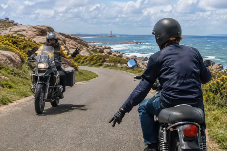 Sur une route côtière bretonne, deux motards qui se croisent se saluent d’un V de la main gauche tendue vers le bas, au milieu des ajoncs en fleur