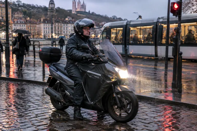 Un homme d’une cinquantaine d’années en tenue de pluie attend au feu sur son scooter 125 équipé d’un tablier et d’un top case, un soir pluvieux à Lyon
