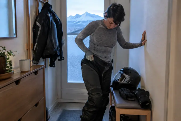 Dans l’entrée d’un appartement à Grenoble, une motarde en sous-couche mérinos et collant thermique enfile son pantalon moto textile avant de partir