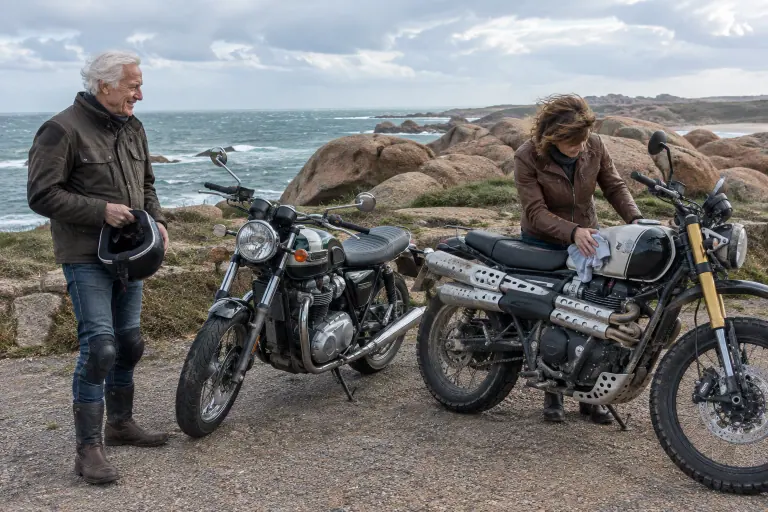 Sur la côte de granit rose en Bretagne, une motarde essuie les embruns sur le réservoir d’un roadster classique anglais pendant que son père sourit