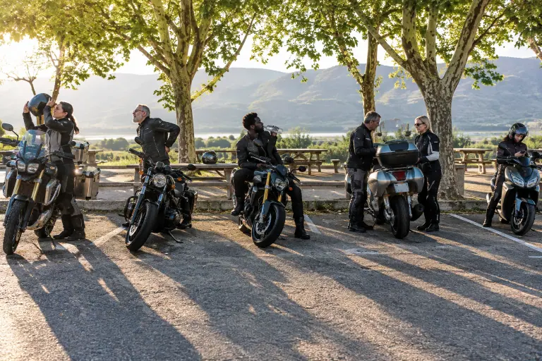 Sur une aire d’autoroute au petit matin, des motards font une pause à côté d’un trail, d’un custom, d’un roadster, d’une routière et d’un scooter