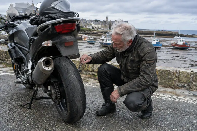 Sur un parking du bord de mer breton, un motard retraité mesure à la jauge la gomme restante sur le pneu arrière usé de sa routière