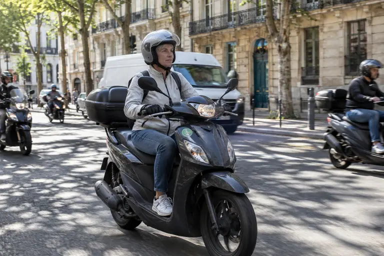 Dans une rue ensoleillée du centre de Marseille, une scootériste aux cheveux gris circule en scooter 125 avec top case parmi la circulation du matin